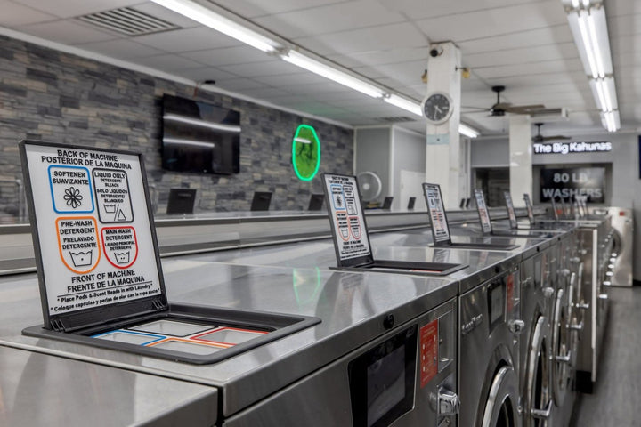 A long row of commercial washers with their top soap dispenser hoppers open, each fitted with a labeled compartment liner, and an 80 LB sign on the wall behind