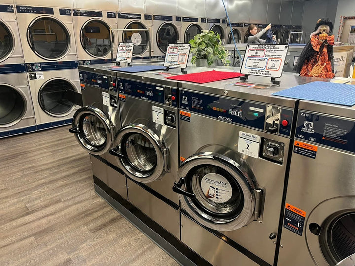 Row of Dexter T-Series front-load washers in a laundromat with numbered decals on the doors and labeled soap hoppers on the top decks