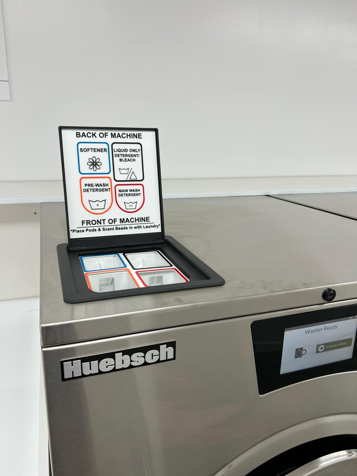 Top deck of a Huebsch Galaxy hardmount washer with the four-compartment supply hopper open and a color-coded liner and label installed.